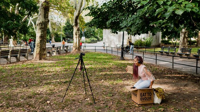 Gahmya Sits In A Park With Signage and Props To Film Her Online Teachings. | Digital Learning