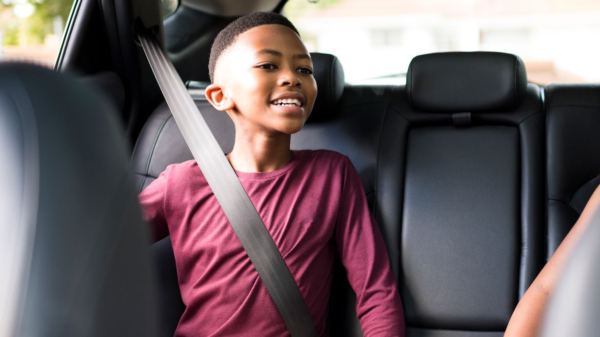 A Young Boy Smiles From The Backseat Of A Vehicle As He Peers Excitedly Into The Drivers Seat. | Teen Drivers